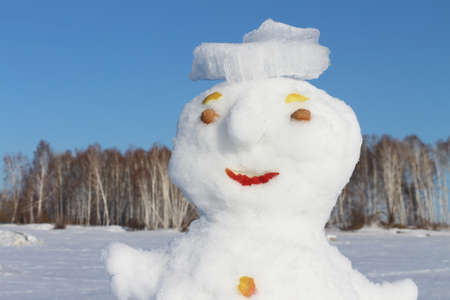 Snowman in a hat from ice standing on a snow glade in park against the blue skyの写真素材