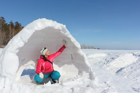 Happy woman in a red jacket sitting in unfinished an igloo   on a snowy glade, Siberia, Russia