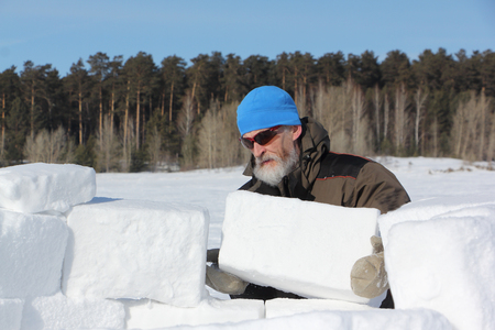 Man in a blue hat and sunglasses building an igloo from snow blocks in the winterの写真素材