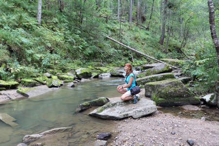 Happy woman with red hair sitting on a stone by the river in summer, Belokurikha River, Belokurikha City, Altai Mountains, Russiaの写真素材