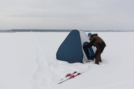 Man in a suit for winter fishing putting up a tent on a frozen river in a blizzard, snowfall and wind, Ob Reservoir, Novosibirsk, Russiaの写真素材