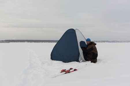 Man in a suit for winter fishing putting up a tent on a frozen river in a blizzard, snowfall and wind, Ob Reservoir, Novosibirsk, Russiaの写真素材