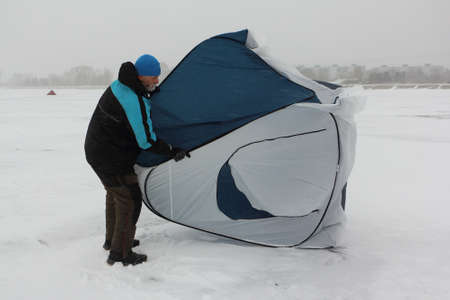 Man in a suit for winter fishing putting up a tent on a frozen river in a blizzard, snowfall and wind, Ob Reservoir, Novosibirsk, Russiaの写真素材