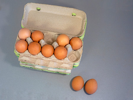 two eggs on a table near brown eggs in a cardboard container on a gray background.
