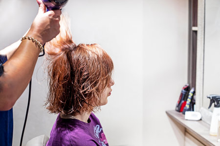 hairdresser drying with a hairdryer in a beauty salon red short hair in a teenager girl on a light background. Everyday life and fashion trendsの写真素材