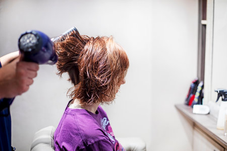 hairdresser makes styling with a hairdryer and a comb in a beauty salon of a teenager girl with red hair. Everyday life and fashion trendsの写真素材