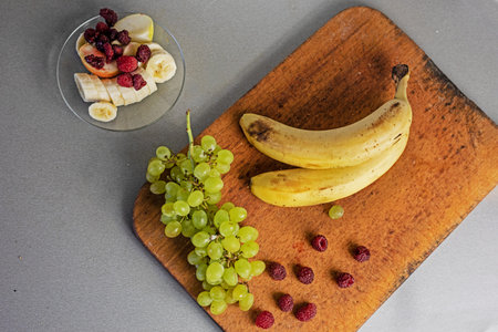 bunch of bananas with a bunch of green grapes and raspberries on a wooden cutting board on a gray backgroundの写真素材