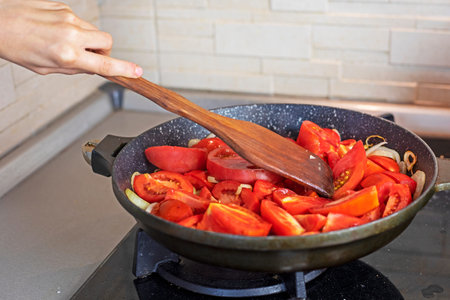 girl stirring tomatoes with onions with a special wooden spatula for cooking in a frying pan in the...