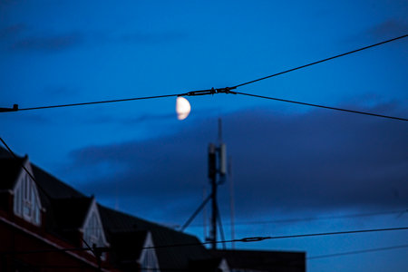 half a moon in the dark sky with clouds in the city above the roofs of houses with wiresの写真素材