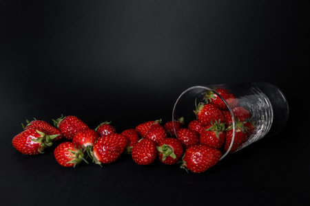 Ripe red strawberries spilling out of clear glass onto dark background in minimalist food...