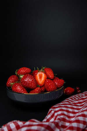 Juicy strawberries served in bowl with white and red gingham napkin over black surface, rustic presentation.の写真素材