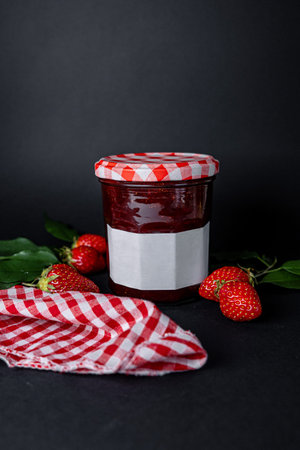 Jar of strawberry jam placed with whole fresh strawberries and green leaves alongside red and white gingham cloth. Cozy and romantic food composition on dark background.の写真素材