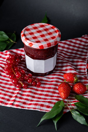 Jar of strawberry jam placed with whole fresh strawberries and green leaves alongside red and white...
