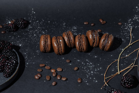 Moody food photography featuring chocolate macarons arranged in row with fresh blackberries and coffee beans on dark textured background.の写真素材