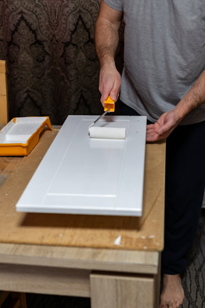 Man painting a white door panel indoors using a roller and tray on a wooden tableの写真素材
