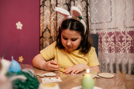 Young girl enjoying craft activity while preparing for Easter with bunny ears and colorful supplies in a cozy roomの写真素材