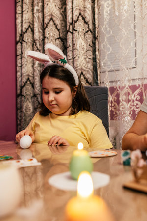 Young girl wearing bunny ears colors Easter eggs at a decorated table in a cozy indoor setting...