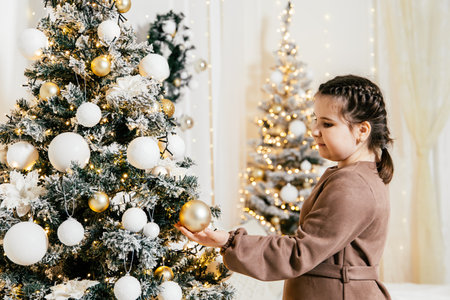 Child decorates Christmas tree with golden ornaments and white decorations in festive indoor settingの写真素材
