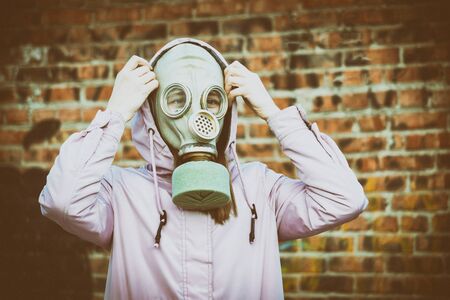 A young girl in a pink jacket, with a gas mask on her face, against a brick wall. Tinted photo with noise overlay, stylized as an old photo. The concept of air pollution, ecology.の写真素材