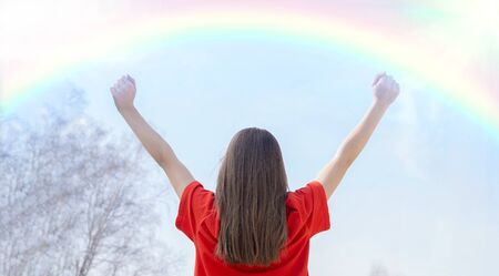 A young girl in a red t-shirt with her hair down, of European nationality,with her hands up, sits with her back to the sky and enjoys the rainbow, blue sky and sun. The concept of spring, joy and hopeの写真素材