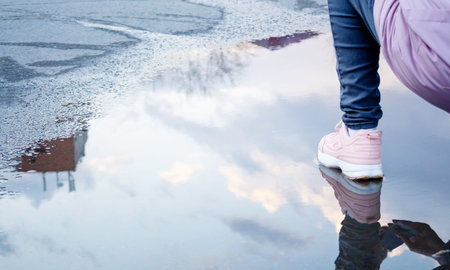 Reflection of women's knees in blue jeans and pink sneakers, blue sky and white clouds in a puddle....