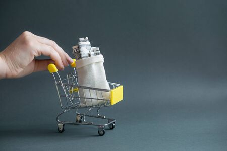A woman's hand holds a yellow shopping cart with a canvas bag filled with money on a dark isolated background. Close-up, copy of the space. The concept of inflationの写真素材