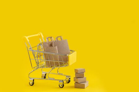 Paper bags in the shopping basket, brown paper boxes next to the basket. The concept of buying or selling goods or services, online purchases. Close-up, yellow isolated backgroundの写真素材