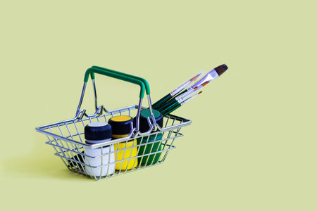Metal shopping basket with green handles, filled with artistic paints: yellow, green and white...