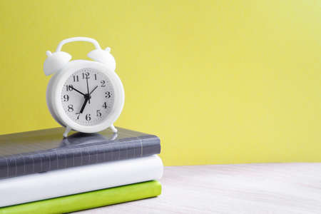 A white alarm clock stands on a stack of three books with colorful spines on the table. Yellow background, close-up, copy space. Back to school conceptの写真素材