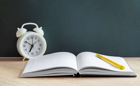 A white alarm clock stands next to an open book with white lined pages and a yellow pencil. Items lie on the background of the school Board. Let's go back to the concept of a school,a copy spaceの写真素材