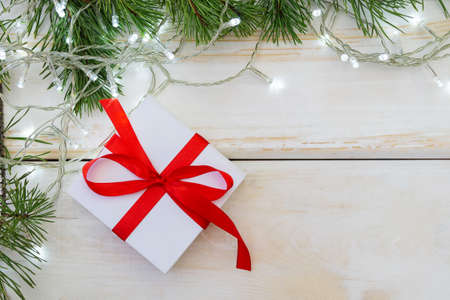 Christmas card. White gift box with a red ribbon on a white wooden background, with pine branches and a glowing garland. Copyspace, flat lay.の写真素材