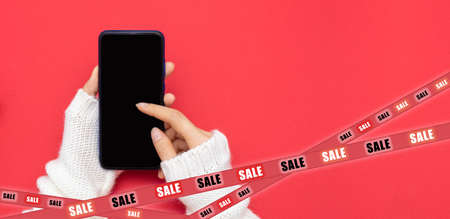 Christmas online shopping . Womens hands in a white sweater place an order on a smartphone on a red background. The inscription on the tapes-sale. Banner, copy of the space. The concept of Black Friの写真素材