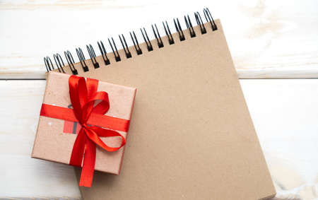 The concept of a wish list for the New year Christmas, a letter to Santa Claus. Brown spiral Notebook, gift box with red bow on white wooden background. Close-up, copy space.の写真素材