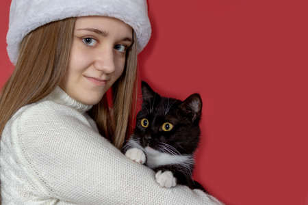A beautiful smiling girl in a red Santa hat and a black cat in her hands on a red isolated backgroundの写真素材