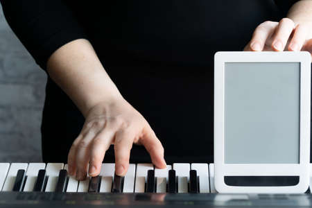 Mock up for online courses play the piano. One hand plays on the keyboard, the other holds the tablet, the screen turned forward. Close-up, selective focus, copy space.の写真素材