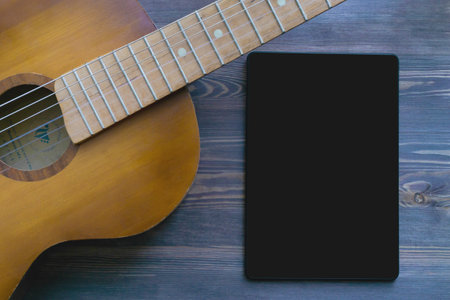 Part of a brown guitar and a vertical screen of a black tablet on a blue wooden surface. The concept of a mock up for advertising online guitar courses.の写真素材