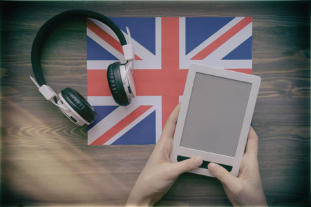 Womens hands hold a tablet against the background of a flag, on which headphones lie. Brown wooden...