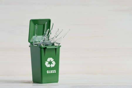 Green container for sorting garbage, with an open lid, glass ampoules and an ecology icon. Close-up, white wooden isolated background. The concept of sorting garbage, waste, for further processing.の写真素材