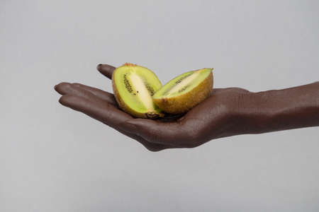 Dark-skinned hand holds a sliced fruit in half, a kiwi. Gray isolated background, close-up, copy spaceの写真素材