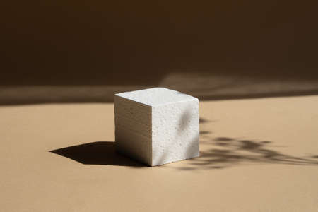 Geometric 3d foam podium for product demonstrations. White cube, in rays of sunlight, casts hard shadow on brown background.の写真素材
