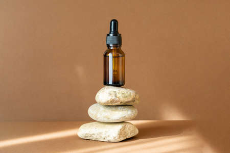 Mock-up of glass dropper bottle stands on improvised stand made of three stones. Rays of sunlight, reflecting from object, fall on beige background. Concept of improvised 3d podium.の写真素材