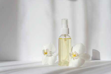 Mock-up of glass bottle with spray bottle surrounded by orchid flowers in rays of sunlight. White background with hard shadows from objects. Concept of skin care, spa treatments, natural cosmetics.の写真素材