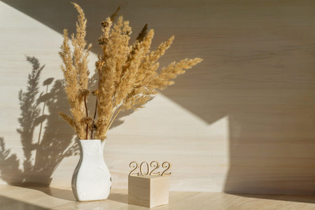 White vase, with dry grass, on beige wooden work table. A 3D podium with numbers 2022, in rays of...