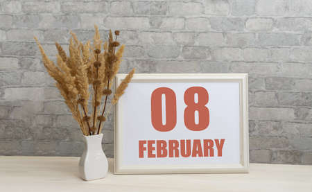 february 8. 8th day of month, calendar date. White vase with ikebana and photo frame with numbers on desktop, opposite brick wall. Concept of day of year, time planner, winter month.の写真素材