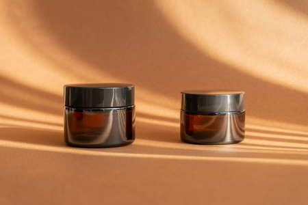 Two jars of amber glass for cosmetics on brown background with shadow in form of stripes. Mock-ups of containers with moisturizer for face, hands, close-up. concept of body care.の写真素材