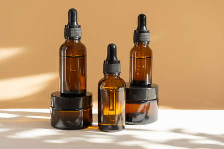 Mock-ups of unmarked cream jars and vials with dropper lids with face serum made of amber glass. Containers for cosmetic products on beige-white background with floral shadow. concept of body care.の写真素材