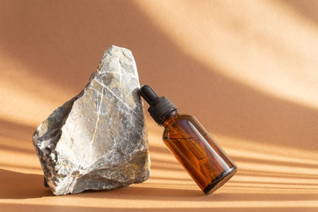 Mock-up of amber-colored glass bottle with dropper lid next to natural granite stone. Facial serum,...