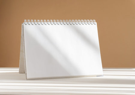 Mockup of clean desktop calendar in rays of sunlight. Unwritten spiral notebook on beige-white...