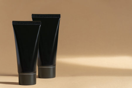 Two dark plastic tubes with cream for face, hands and body on beige background with abstract shadow....