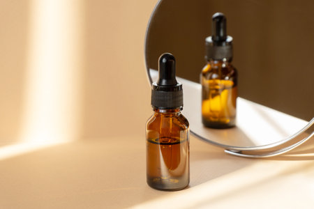 Cosmetic serum for face in glass bottle on beige background in rays of sunlight. Dry skin care product, essential oil for moisturizing skin is reflected in mirror. Copy space, close-upの写真素材